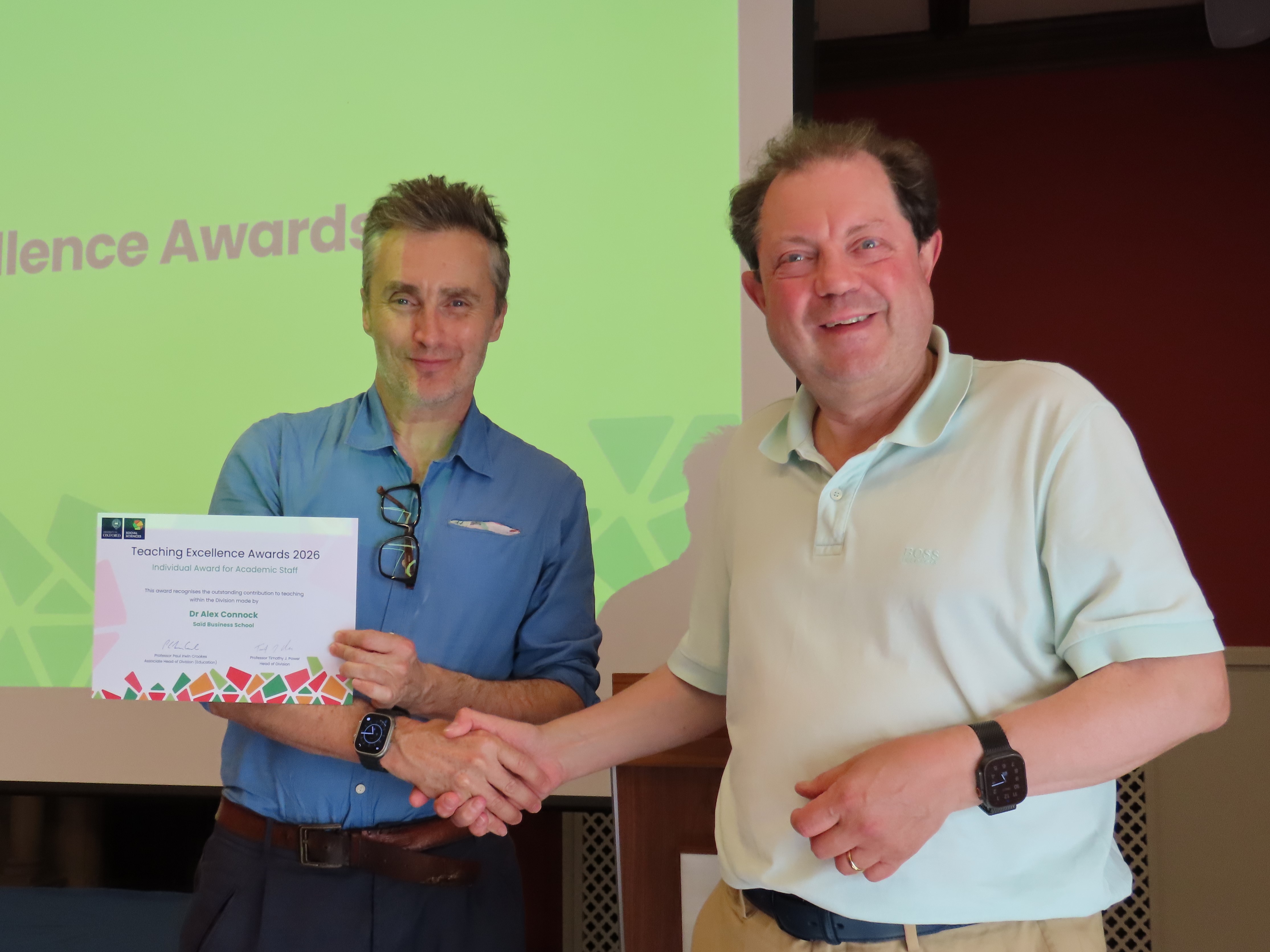 Dr Alex Connock, wearing a blue shirt and holding a certificate, shakes hands with Professor Paul Irwin Crookes