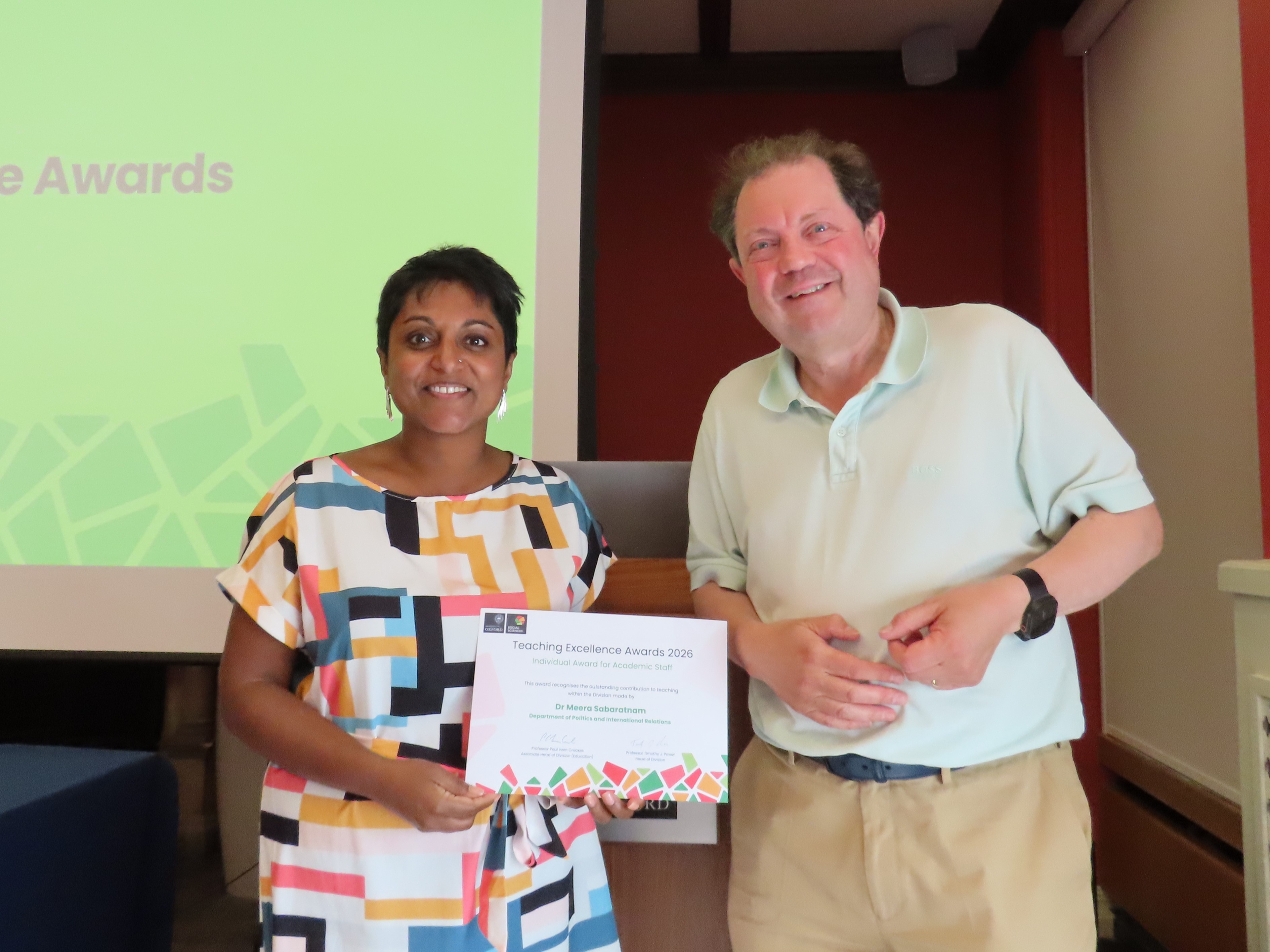 Dr Meera Sabaratnam, wearing a summer dress with a bright abstract pattern, holds a certificate next to Professor Paul Irwin Crookes
