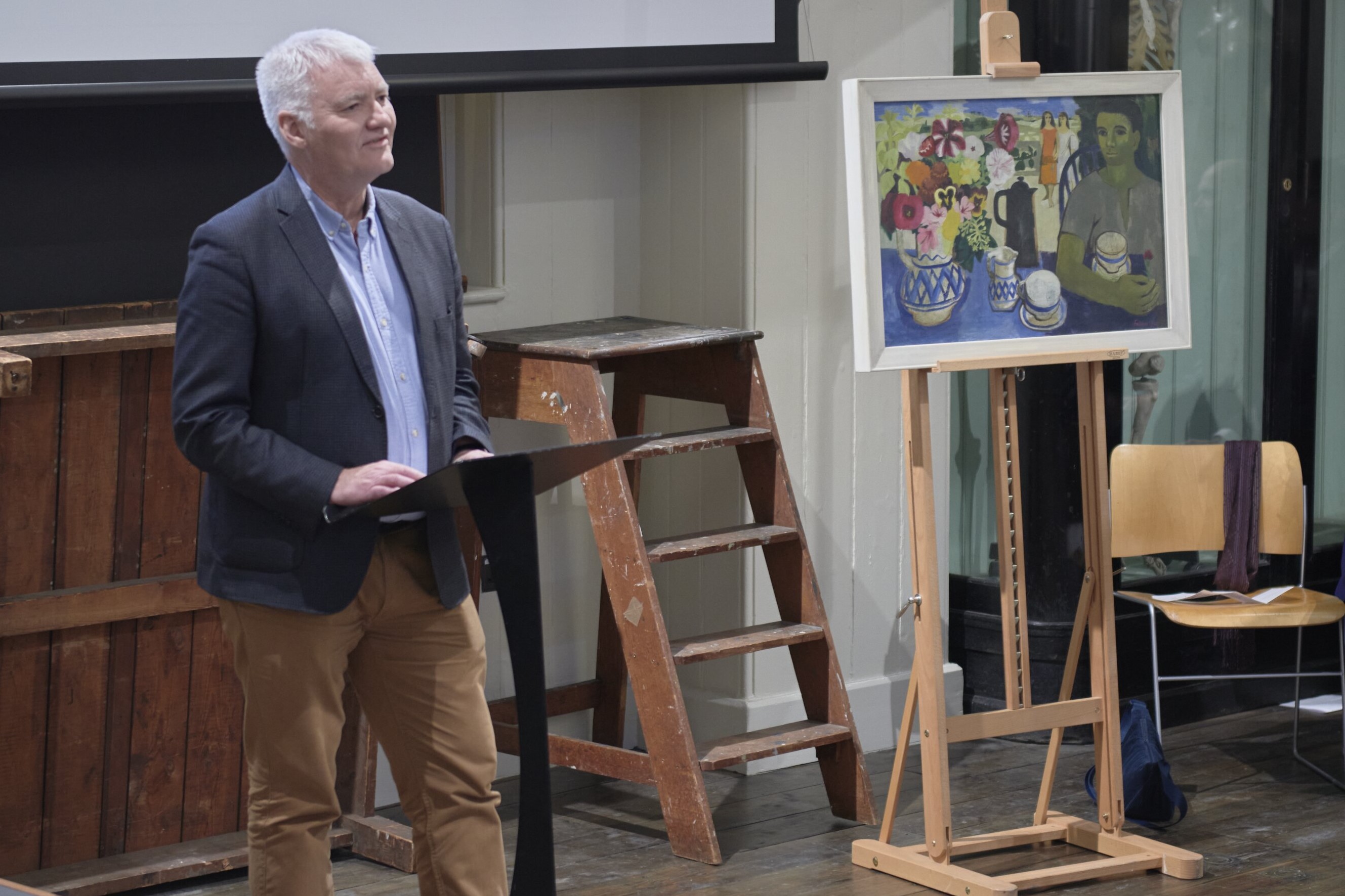 Associate Professor Ian Thompson is standing in front of a ladder and some art. He is wearing brown trousers, blue shirt and navy blazer and presenting to the room. 