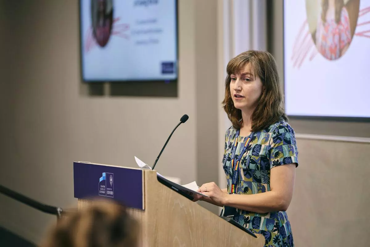 White woman with shoulder-length hair and a floral pattern dress addresses the audience from a lectern