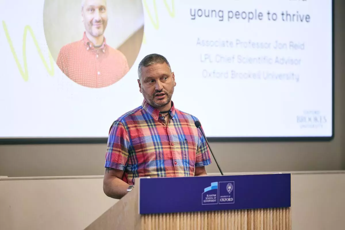 White man with short hair and a checked pattern shirt addresses the audience from a lectern