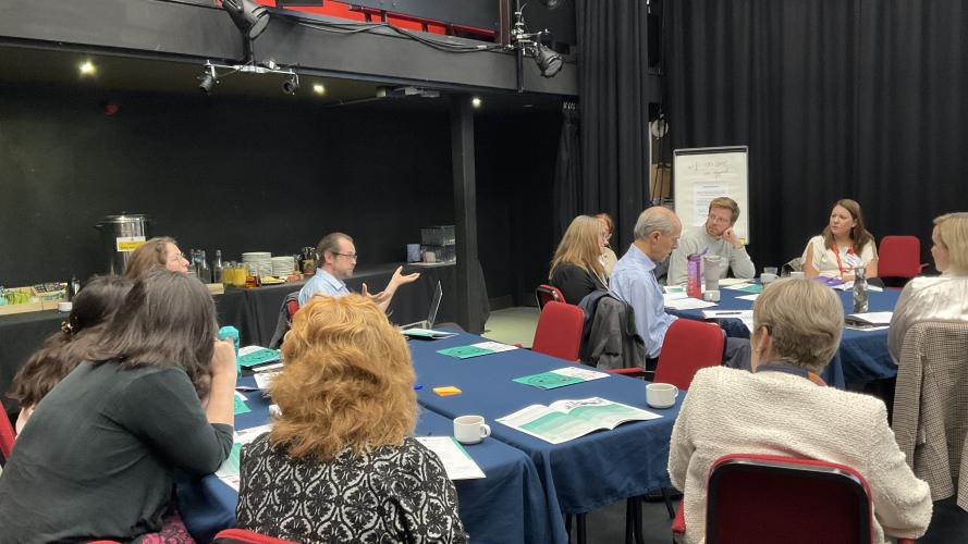 Participants seated around conference tables engage in a workshop discussion, with one individual presenting to the group.