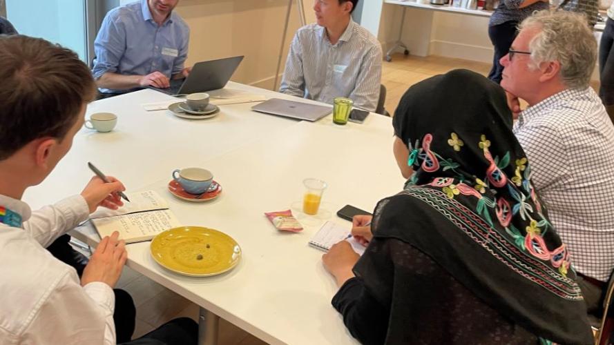 Researchers seated around a table are in conversation; one is typing, two others are taking notes by hand. In the background is a flipchart and more people in conversation