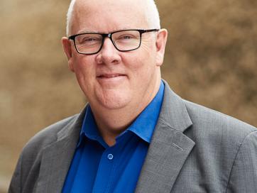 Tim Power wears a blue shirt and grey jacket. He is white and has white hair, he wears glasses with black rims. He is standing outside with a brick wall behind him, which is blurred. He is smiling.