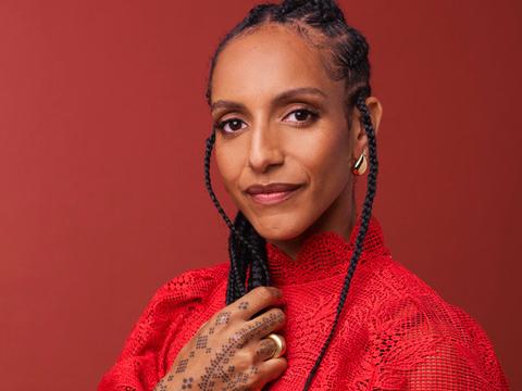 Portrait of Afua Hirsch, a Black woman with long braids, wearing a red lace top and gold jewellery, posing confidently against a warm red background. Her hand, adorned with a dotted design, rests over her chest.