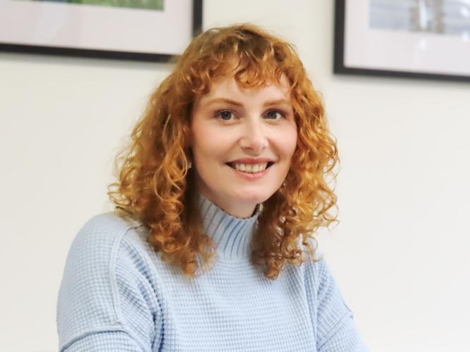 kate oconnor, ha shoulder length red hair, wearing a blue top smiling at the camera.
