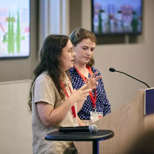 Two young women address the audience from a lectern, both have long brown hair and one wears a grey top while the other wears a blue patterned top