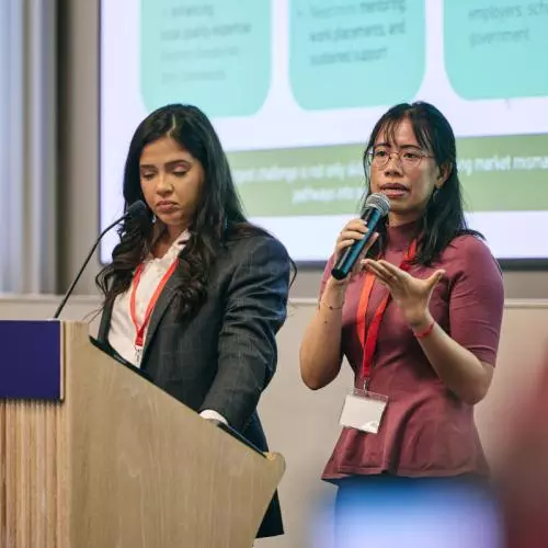 Two women address the audience from a lectern, one holding a microphone