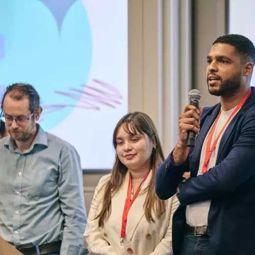 Three people stand in front of an audience, one is speaking and holding a microphone