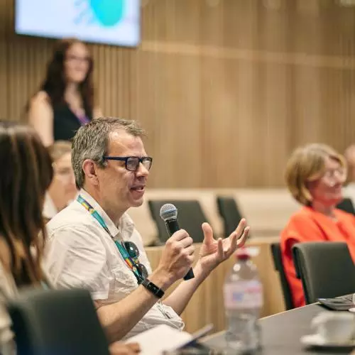 A white man with glasses in the audience asks a question holding a microphone