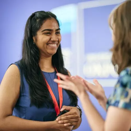 Two women speaking, one in the foreground is blurred and the other has long dark hair and a blue top