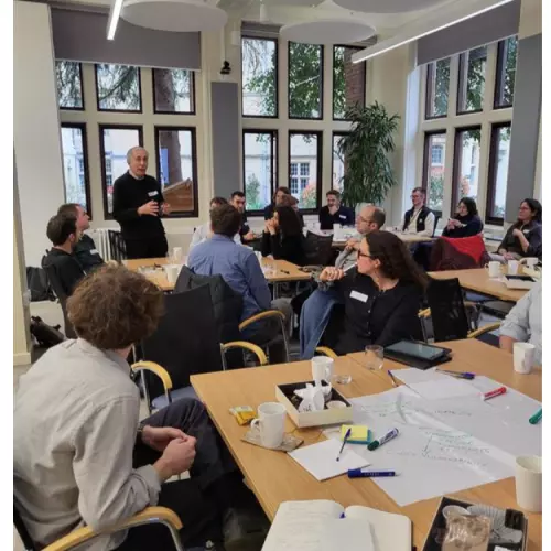 A male academic addresses a room full of researchers who listening to him attentively, and seated around tables that are covered in pens and paper, brainstorming activities, and lunch