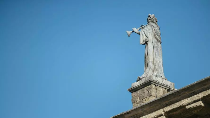 A statue of a robed figure blowing a trumpet, positioned on the roof of a building against a bright blue sky