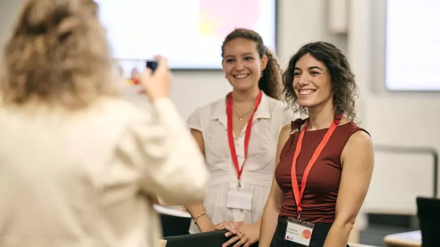Two women stand posing for a photograph, one is wearing a white top and the other a burgundy top, both have dark wavy hair