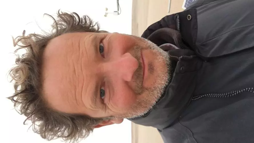Giles has dark hair and is smiling into the camera