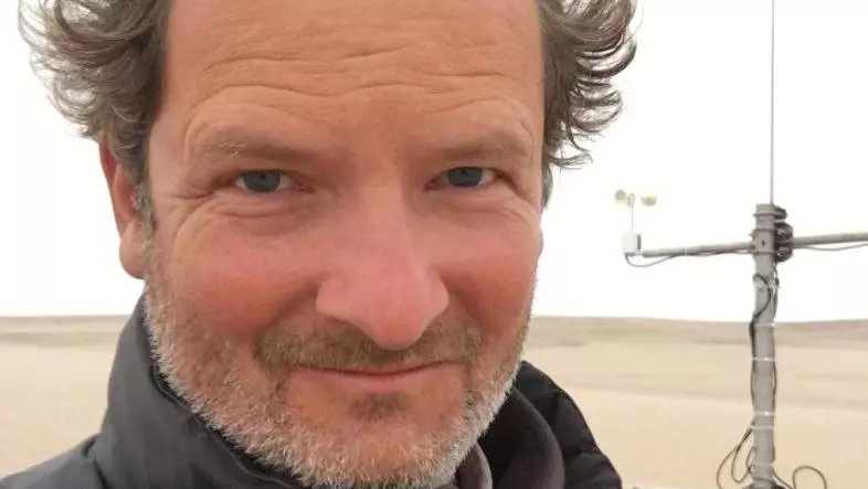 Giles Wiggs has short curly brown hair. He is wearing a black jacket and standing outside