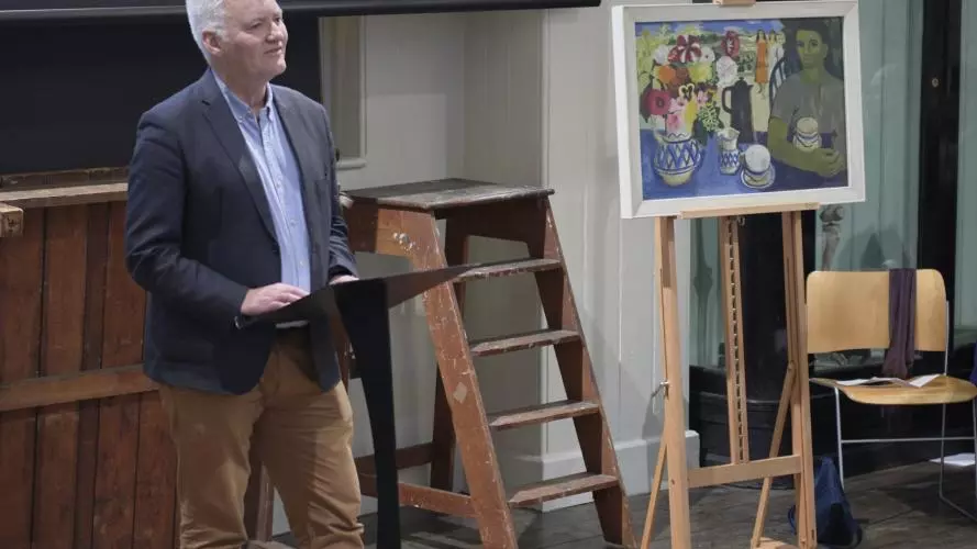 Associate Professor Ian Thompson is standing in front of a ladder and some art. He is wearing brown trousers, blue shirt and navy blazer and presenting to the room. 