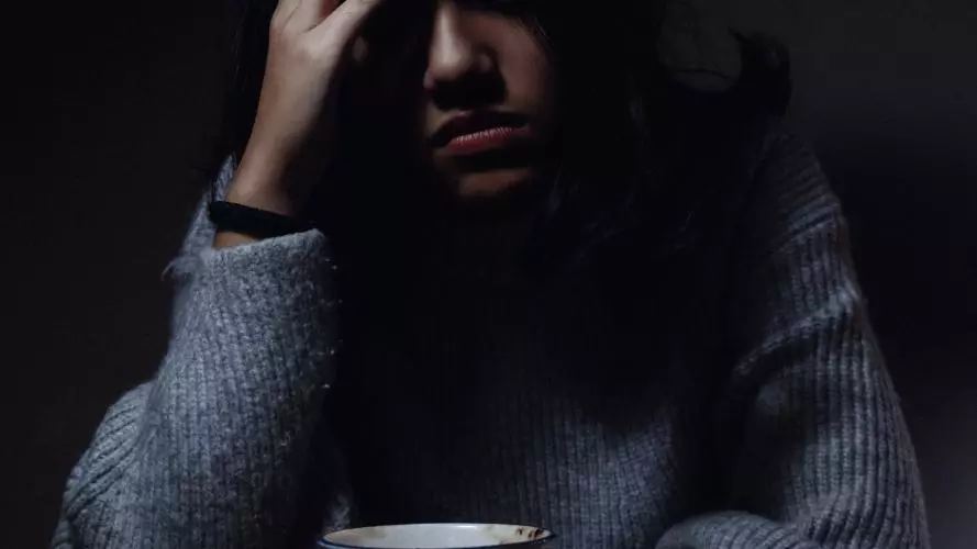 A young woman sits in a dark room with her head in one hand, leaning over a cup of tea