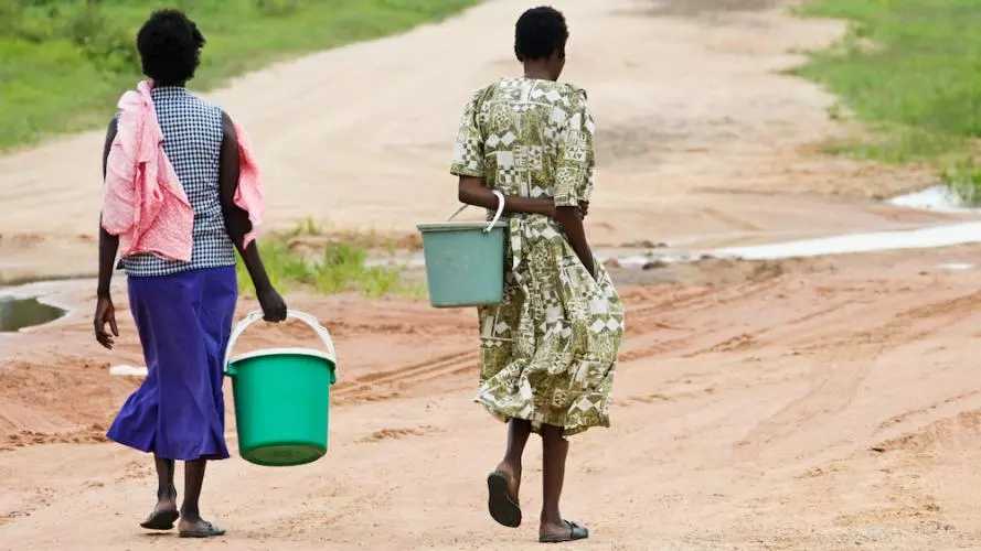 The view from behind of two women from an African village carrying empty buckets to collect water. The ground is mostly arid and dry, and there is grass in the distance.