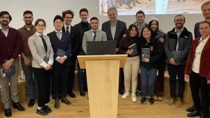 Seventeen smiling, smartly dressed people stand behind a lectern, some holding copies of a book. 