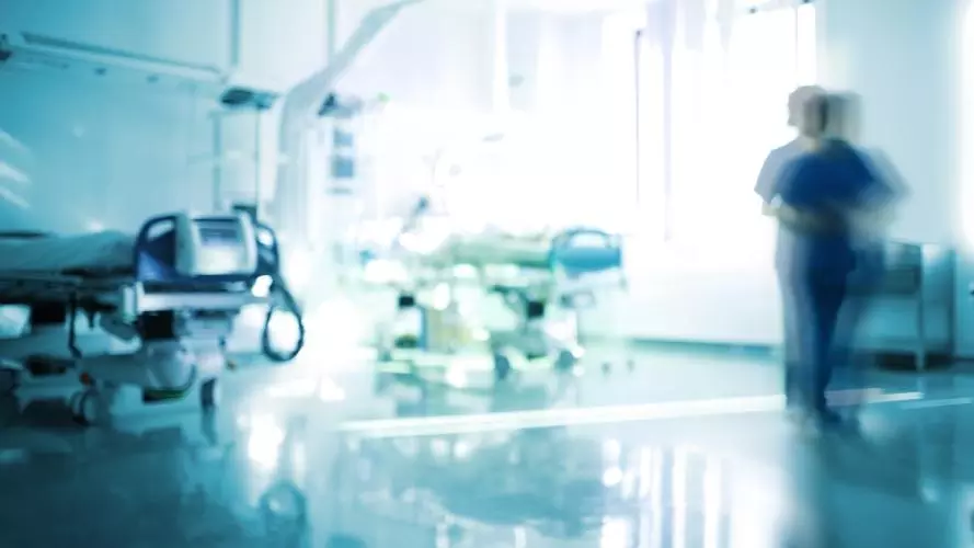 Blurred image of a hospital setting, showing two hospital beds and a medical professional walking past