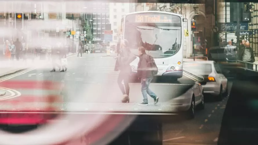 Photo of a bus window reflecting people walking and a bus and cars driving through a street. The image is slightly blurred and gives the impression of movement.