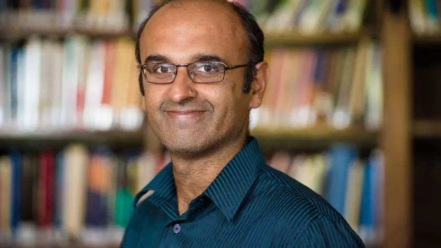 Portrait photo of Professor Yadvinder Malhi, smiling against a blurred background of shelves full of books