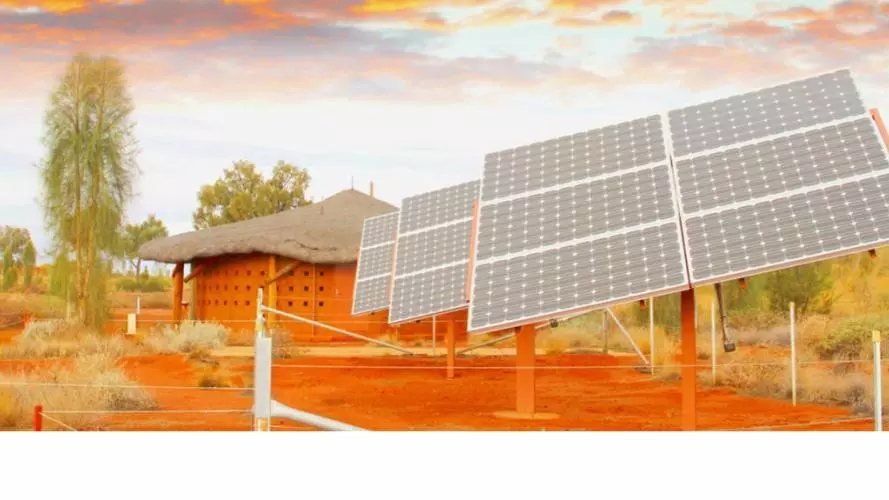 An arid landscape with a thatch-roofed mud hut in the background, in front of which are constructed three large, shiny solar panels, surrounded by fencing
