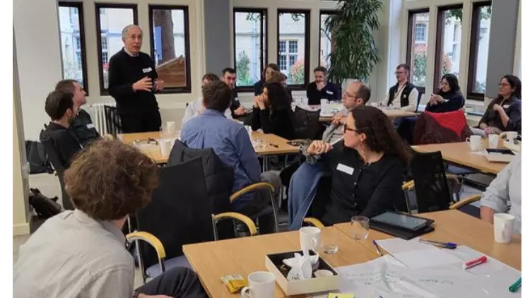A male academic addresses a room full of researchers who listening to him attentively, and seated around tables that are covered in pens and paper, brainstorming activities, and lunch