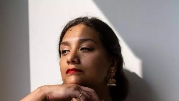 Dr. Shyama Vermeersch is wearing striking red lipstick and is looking off to the side. She is also wearing a green scarf and has dark brown hair tied back.