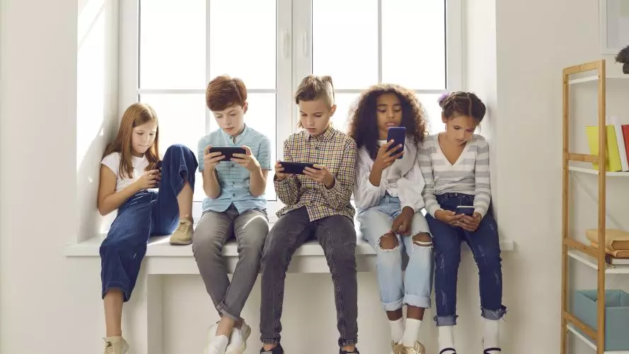 Group of children of different nationalities sit in a row on the windowsill and with lack of emotion play online games or read social networks on mobile phones