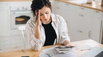 A woman with dark hair sits at a desk, wearing a black tshirt and white overshirt. She looks stressed, resting her head on one hand, while looking at piles of receipts next to a calculator. Credit: sheremetio - stock.adobe.com