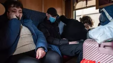 Three people in a train carriage surrounded by luggage. Two look solemn, the other faces the window with their head in their hand.
