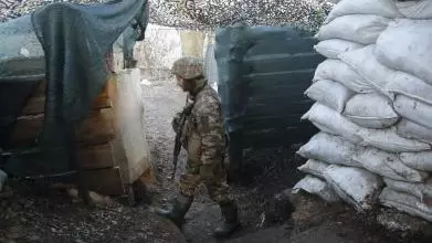 A Ukrainian soldier dressed in camouflage uniform and holding a gun pointed towards the ground patrols a post fortified with sandbags and draped in camouflage.