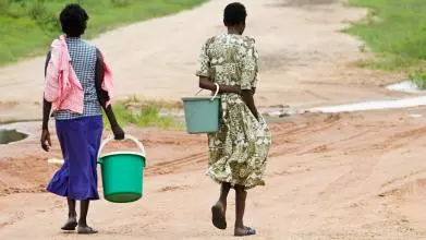 The view from behind of two women from an African village carrying empty buckets to collect water. The ground is mostly arid and dry, and there is grass in the distance.