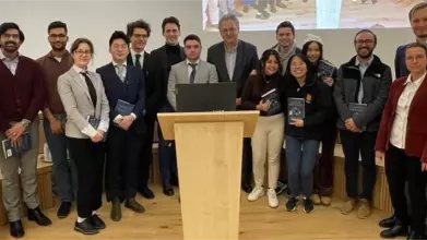 Seventeen smiling, smartly dressed people stand behind a lectern, some holding copies of a book. 