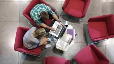 Two students look at a laptop, viewed from above so we can't see their faces. They are sitting on red chairs, with 4 empty chairs and a table in the middle covered in books.