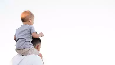 A young boy sits on his father's shoulders. The father is holding the boy's ankles to stabilise him while the boy has his hands in the air.