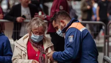 A woman wearing a mask in a queue of people presents vaccine documentation to an official permitting entry