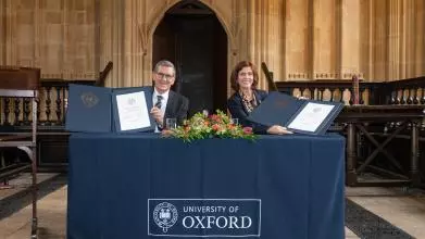 Professor Louise Richardson (VC of University of Oxford) and Professor Reinhold Geilsdoerfer (MD of the Dieter Schwarz Foundation) sit at an Oxford-blue branded desk hoding signed agreements and smiling