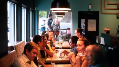 View from inside a pub restaurant, showing three tables surrounded by groups of friends smiling, laughing, talking, and enjoying drinks.