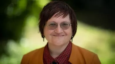 Professor Erduran has short dark hair and round glasses. She is wearing a black and red checked blouse and orange blazer. 