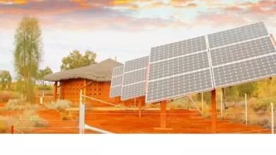 An arid landscape with a thatch-roofed mud hut in the background, in front of which are constructed three large, shiny solar panels, surrounded by fencing