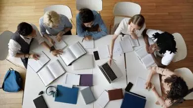 A group of six students sitting in a semi-circle around a white table, which is covered in books and laptops. 