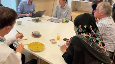 Researchers seated around a table are in conversation; one is typing, two others are taking notes by hand. In the background is a flipchart and more people in conversation