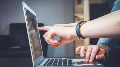 A person points to something on a computer screen to guide the computer user, whose fingers are moving the mouse