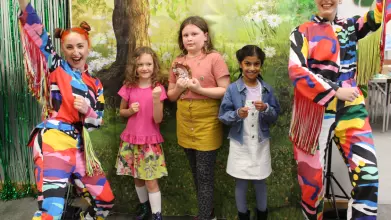 Two adults, dressed as mutli-coloured, cheering butterflies, and three smiling children, pose  as "superpillars" in front of a forest backdrop.