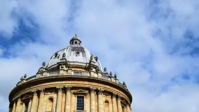 A view of the circular Radcliffe Camera library showing the blue-grey leaded roof and yellow standstone walls against a bright blue sky with whispy clouds