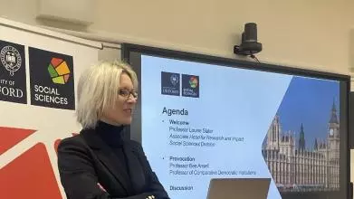 Professor Louise Slate stands at a lecture podium in front of Social Sciences Divisional branding and a slide of the agenda for the Democracy seminar