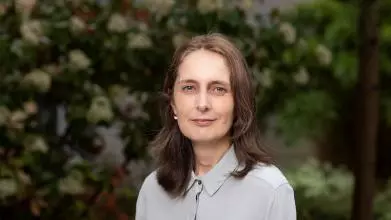 Professor Rebecca Eynon stands in front of some foliage. She has shoulder-length brown hair and is wearing a light grey blouse. 